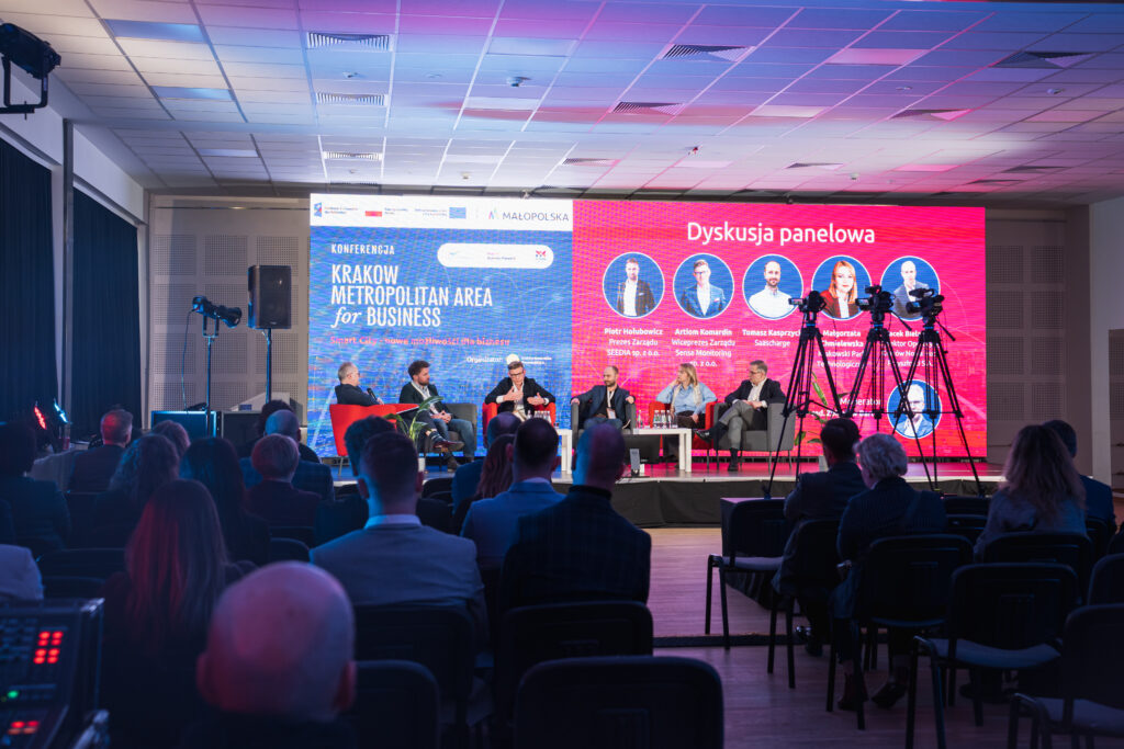 Grupa uczestników siedzi na widowni podczas konferencji „Krakow Metropolitan Area for Business. Smart City - nowe możliwości dla biznesu”, skupiona na prezentacji odbywającej się na scenie